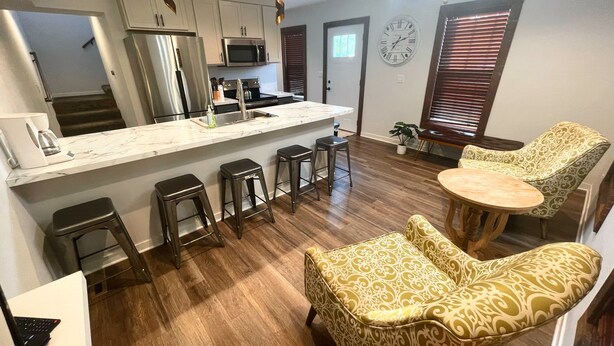 A modern kitchen with stainless steel appliances, a marble countertop, and a central island with bar stools. There are two patterned chairs and a small round table.