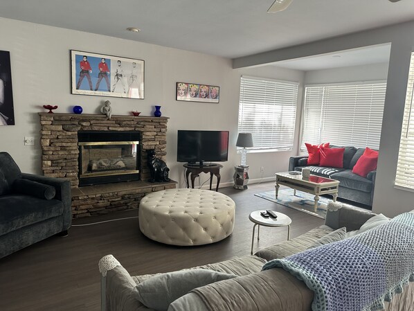 A living room with a stone fireplace, a flat-screen TV, a grey sofa with red cushions, and a grey armchair.