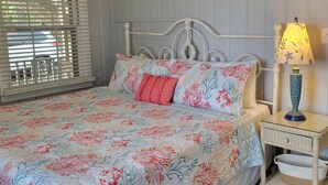 Une chambre à coucher avec une couvre-lit à motifs floraux, une tête de lit en métal blanc, une lampe de chevet et une table de chevet en osier.
