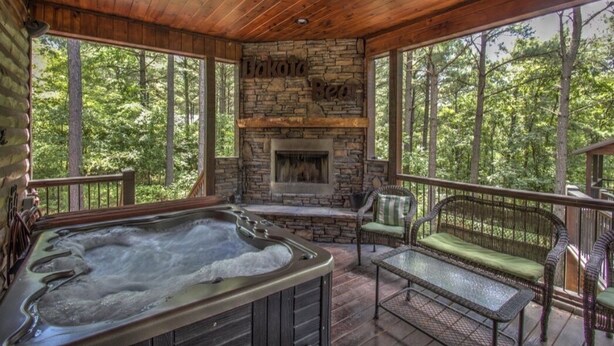 A wooden deck with a stone fireplace, a hot tub, wicker furniture, and a view of the forest.