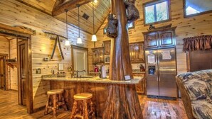 A rustic cabin interior with a wooden bar, stools, a refrigerator, and a bed.