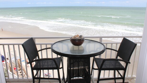 Een balkon met een ronde tafel en twee stoelen, met uitzicht op een strand en golven.