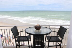 A balcony with a round table and two chairs, overlooking a beach with waves.