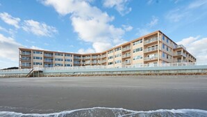 Hotel aan het strand met meerdere balkons, een zandstrand op de voorgrond en een heldere lucht.