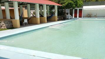 A swimming pool with a tiled deck, a covered seating area with chairs, and a palm tree in the background.