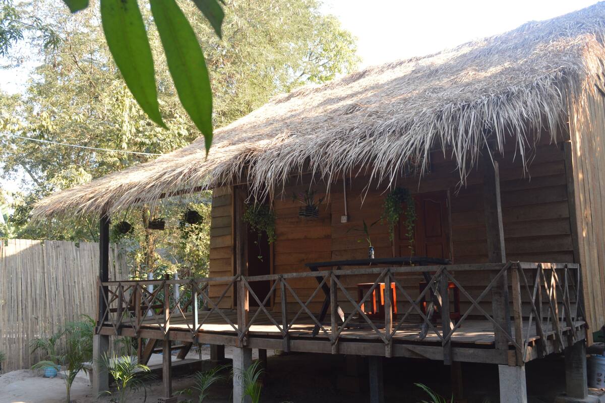 A wooden cabin with a thatched roof, a wooden deck, and hanging plants.