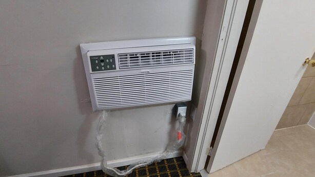 A white portable air conditioner mounted on a wall with a control panel, a power outlet, and a plastic bag hanging from the bottom. - Boone Trail Inn (Middlesboro)
