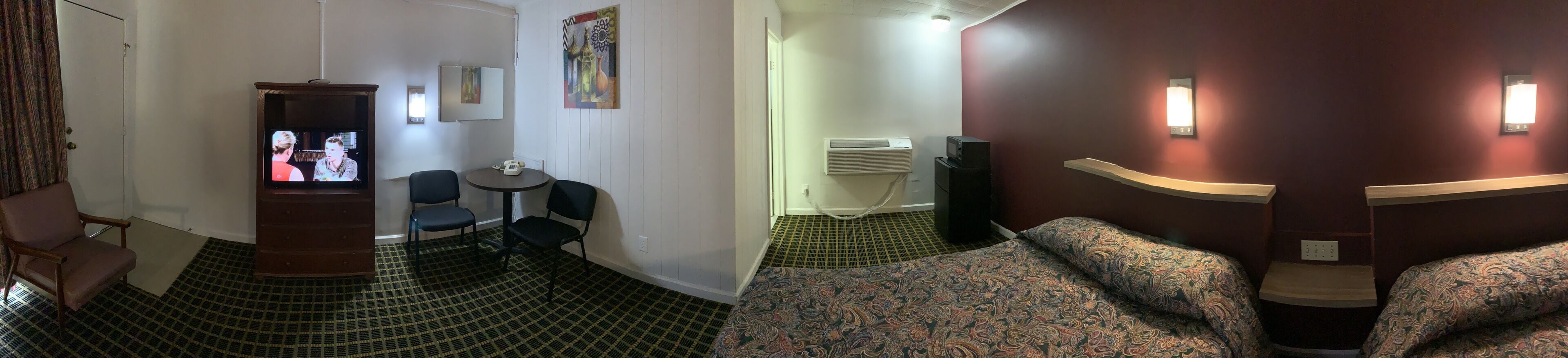Habitación de hotel con televisión, una pequeña mesa con sillas, alfombra estampada, cama con cubrecama estampado y aire acondicionado instalado en la pared.
