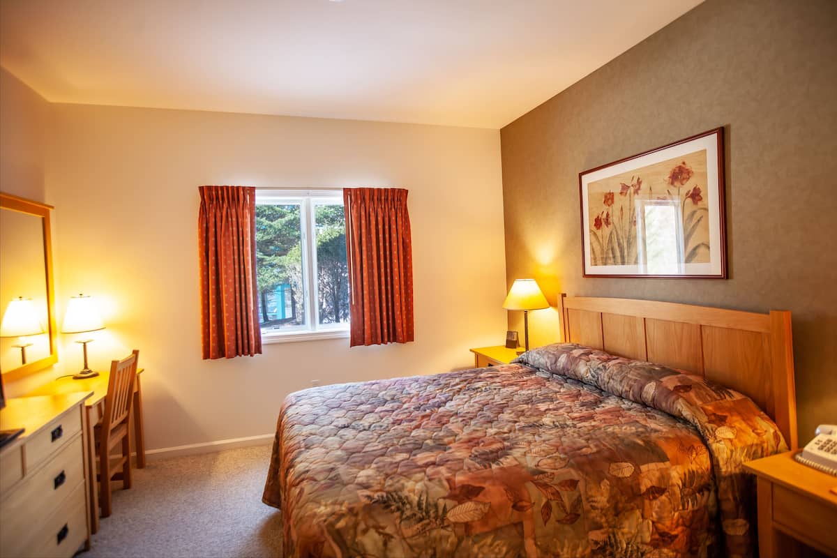 A bedroom with a bed, two bedside tables, a nightstand, a desk with a chair, a window with curtains, and a framed picture on the wall.