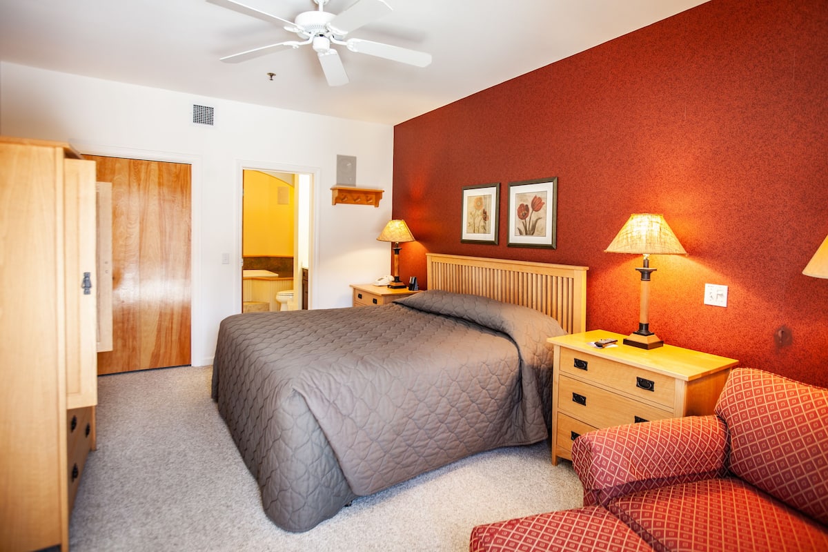 A bedroom with a bed, bedside tables, a nightstand, a chair, a ceiling fan, and two wall-mounted lamps.
