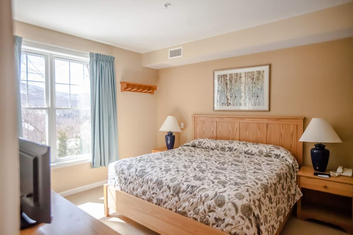 A bedroom with a bed, two bedside tables with lamps, a mounted TV, a window with curtains, and a framed picture on the wall.