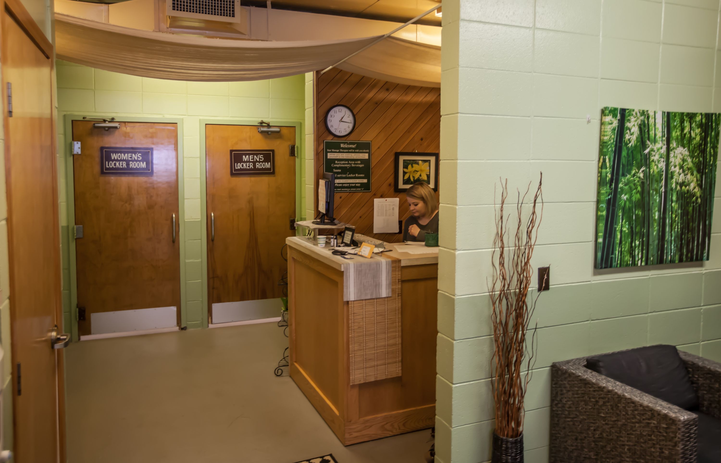 A reception area with a wooden reception desk, a clock, and a welcome sign. There are two doors labeled "Womens Locker Room" and "Mens Locker Room." A chair and a vase of branches are visible in the foreground.
