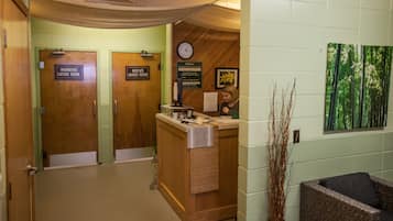 A reception area with a wooden reception desk, a clock, and a welcome sign. There are two doors labeled "Womens Locker Room" and "Mens Locker Room." A chair and a vase of branches are visible in the foreground.