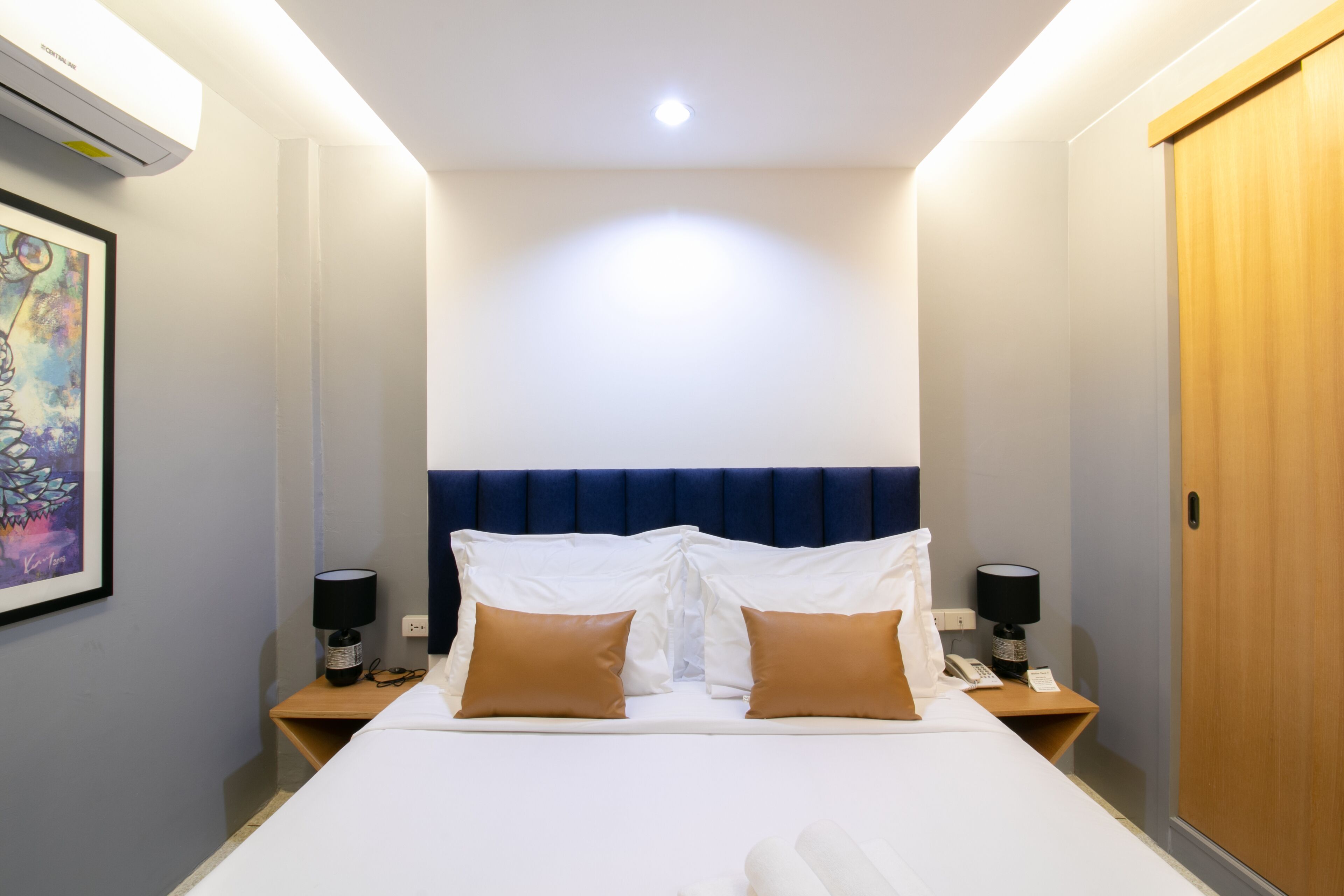 A hotel room with a large bed, two bedside tables, a wall-mounted air conditioner, and a wooden door.