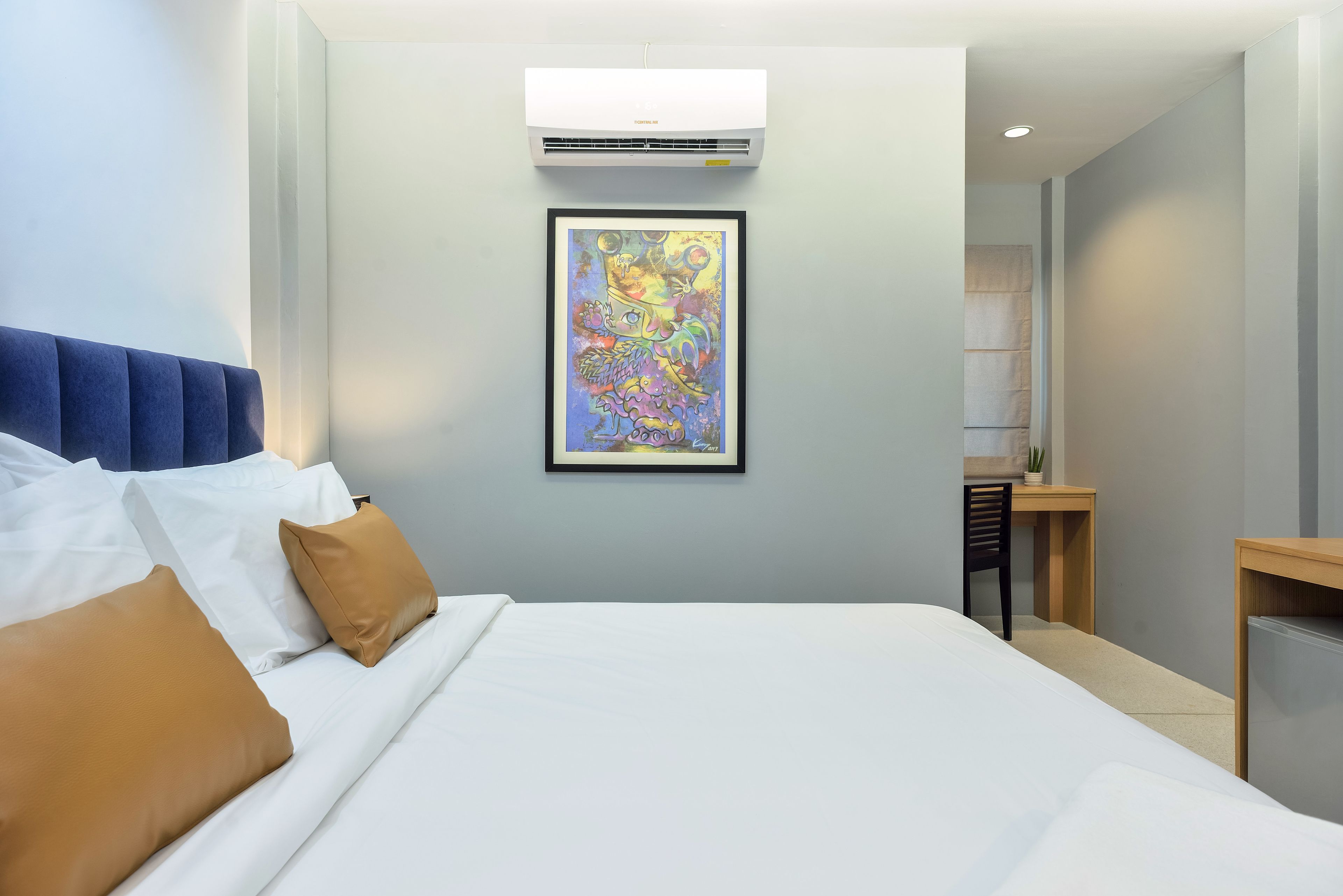 A hotel room with a bed, a desk, a chair, an air conditioner, and a framed artwork on the wall.