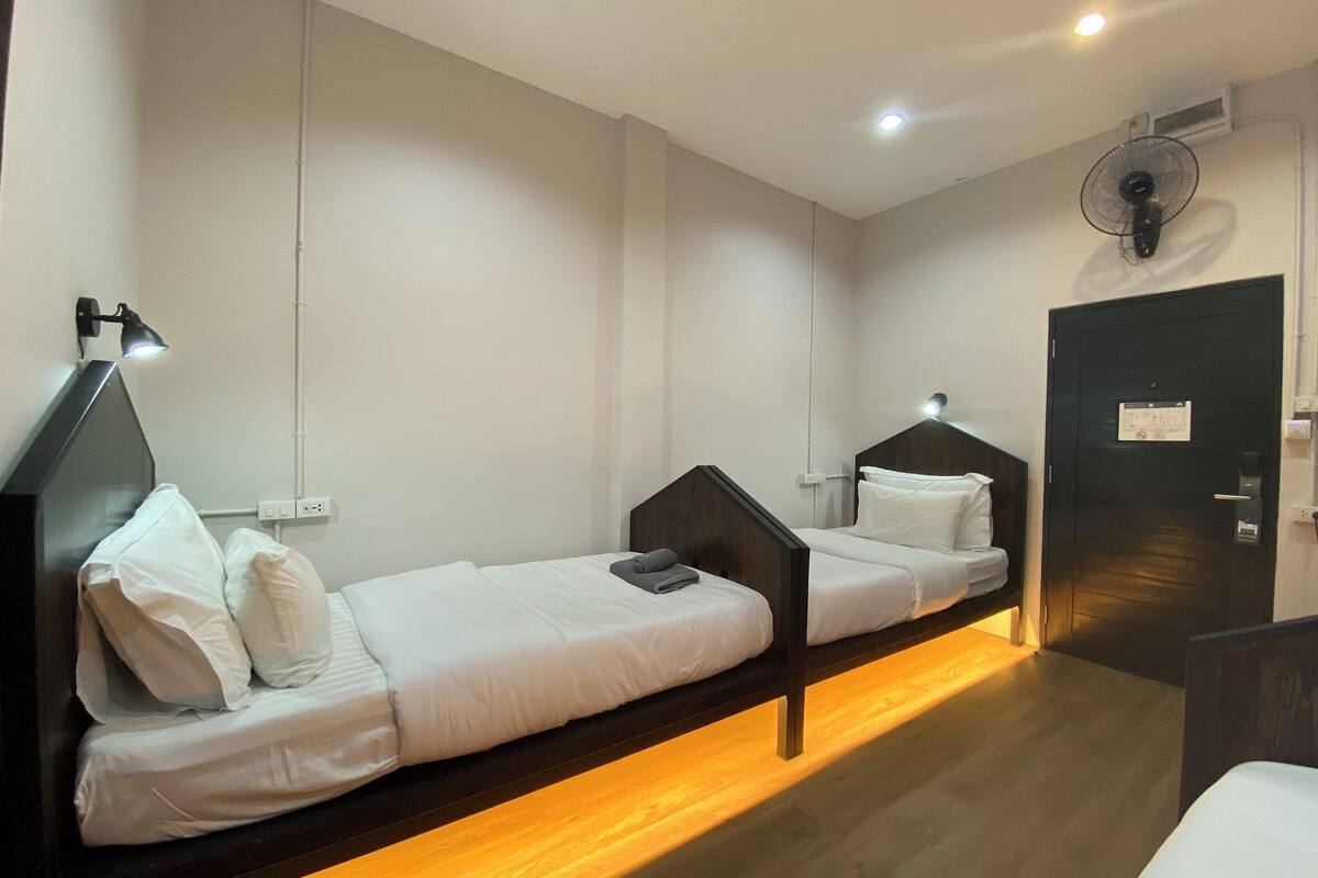 A hotel room with two single beds, a desk, a chair, a standing fan, and a door with a sign.