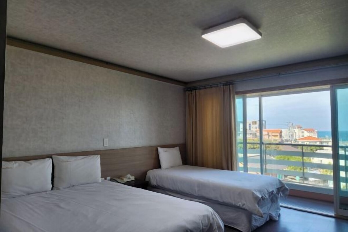 A hotel room with two beds, a large window with a view of buildings and the sea, and a ceiling light.