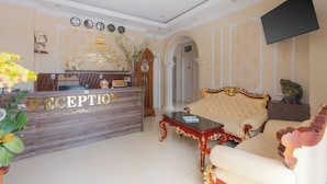 A reception area with a wooden reception desk, a sofa, and a television mounted on the wall. - Green Meadow Central Đà Lạt - O'TEL (Xuan Huong - Da Lat)
