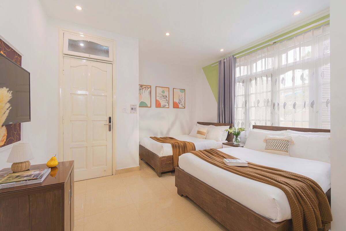 A hotel room with two beds, a desk with a lamp, a door, and a window with sheer curtains.