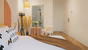A hotel room with a bed, a bedside table with a lamp, a chair, a small table with flowers, and a shower area. - Green Meadow Central Đà Lạt - O'TEL (Xuan Huong - Da Lat)