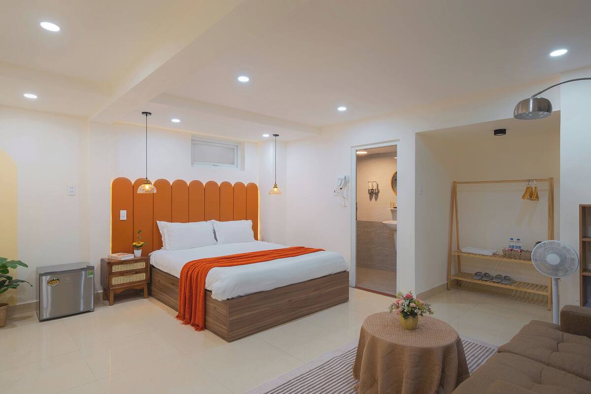 A modern bedroom with a large bed, bedside tables, a small refrigerator, a fan, and a mirror.