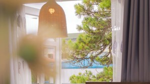 A bedroom with a bed, a bedside table with a vase of flowers, a hanging light fixture, and a view of a balcony with greenery and a building in the background. - Green Meadow Central Đà Lạt - O'TEL (Xuan Huong - Da Lat)