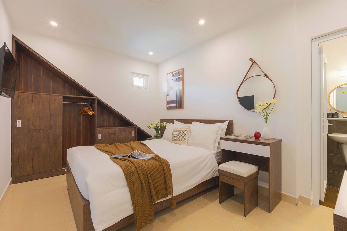A bedroom with a bed, bedside table, bench, wooden wardrobe, and a bathroom with a sink and mirror.