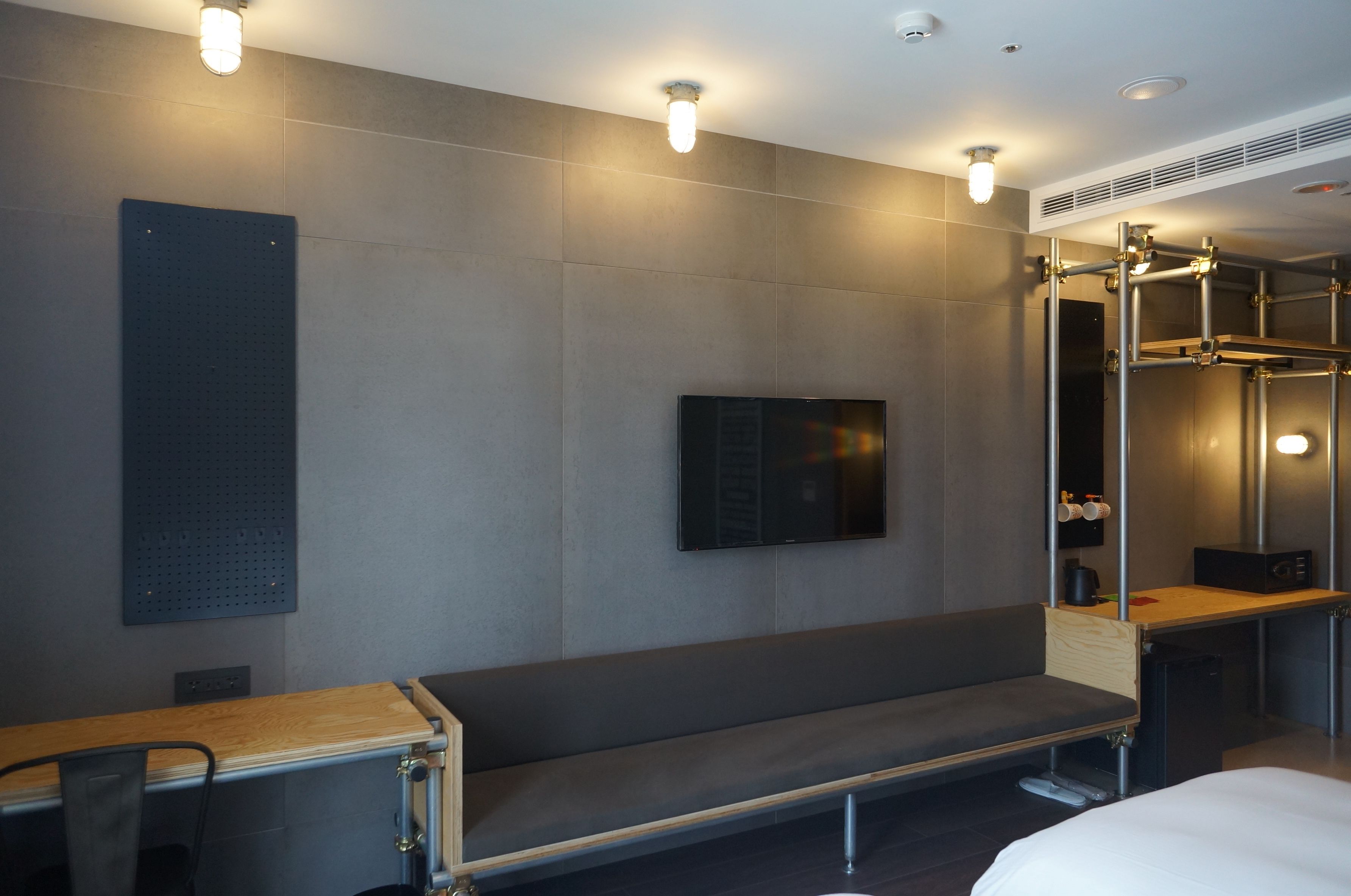 A modern hotel room with a large bed, a wooden bench, a TV mounted on the wall, and a shower area with a glass door.