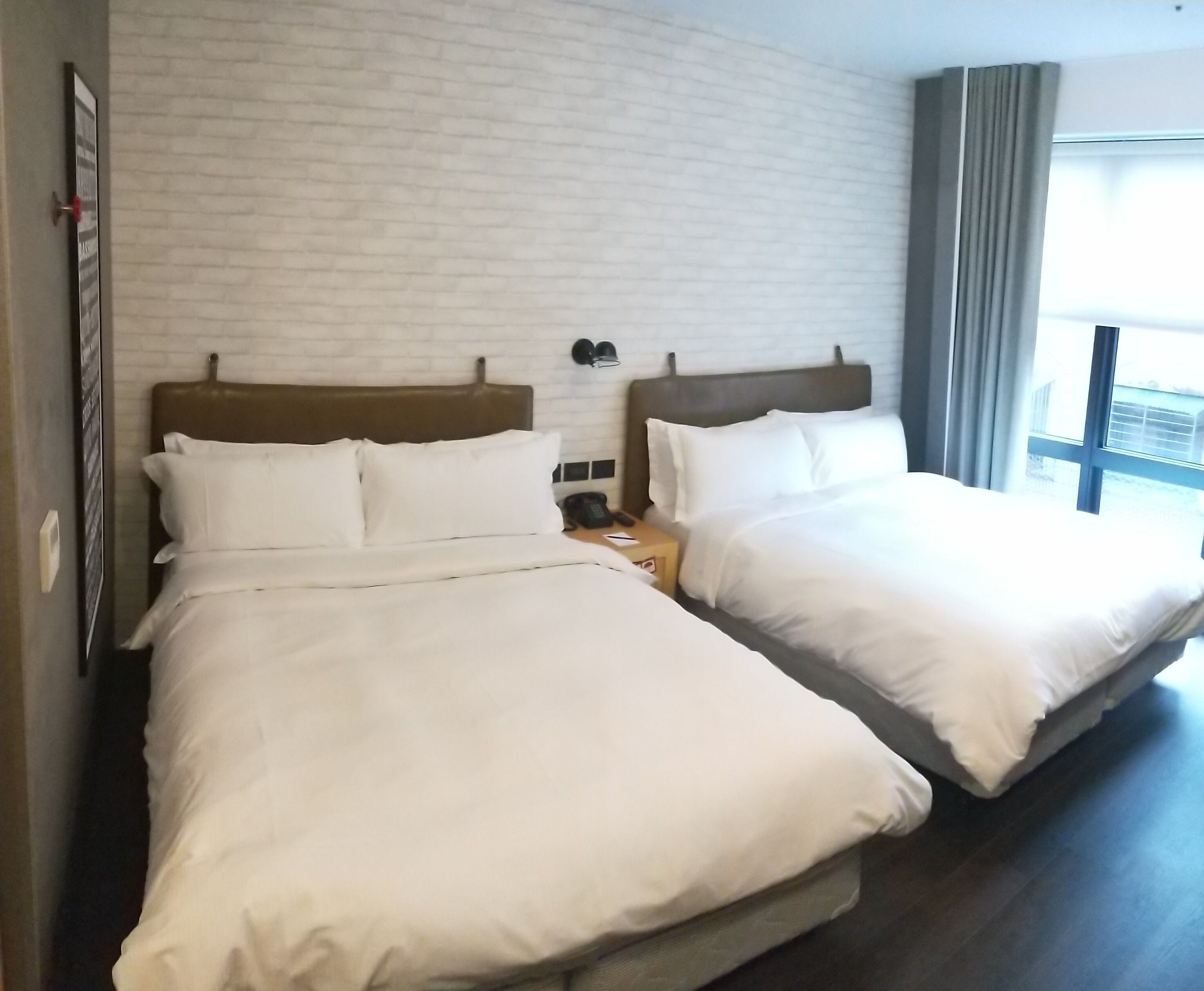 Two twin beds with white linens, a brick-patterned wall, and a window with curtains.