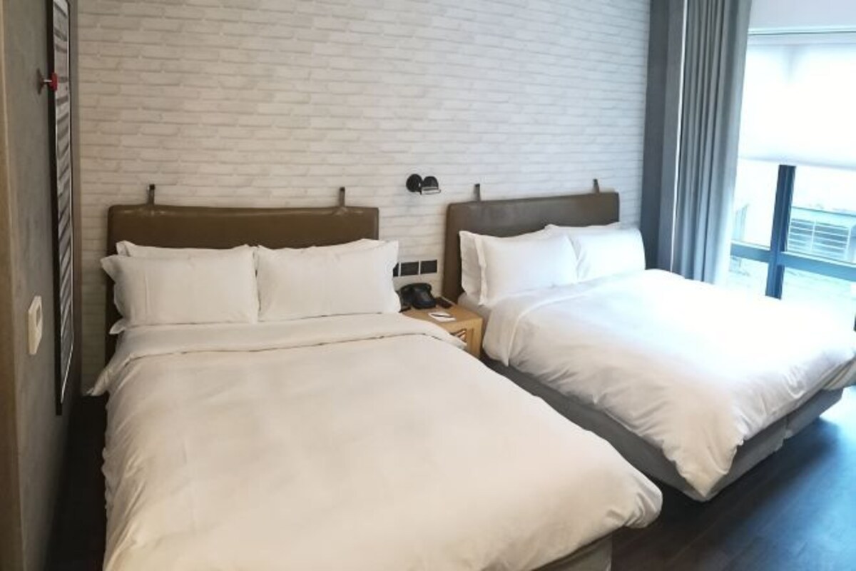 Habitación de hotel con dos camas, una pared de ladrillo, una mesita de noche con un teléfono y una ventana con vistas.