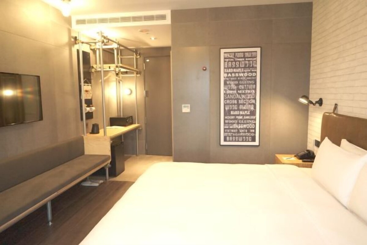 A hotel room with a bed, a bench, a TV, a mirror, a wall-mounted light, and a poster.