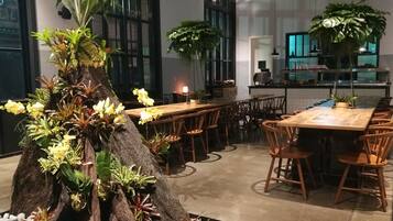 A restaurant with wooden tables and chairs, a large tree centerpiece, and a kitchen in the background.