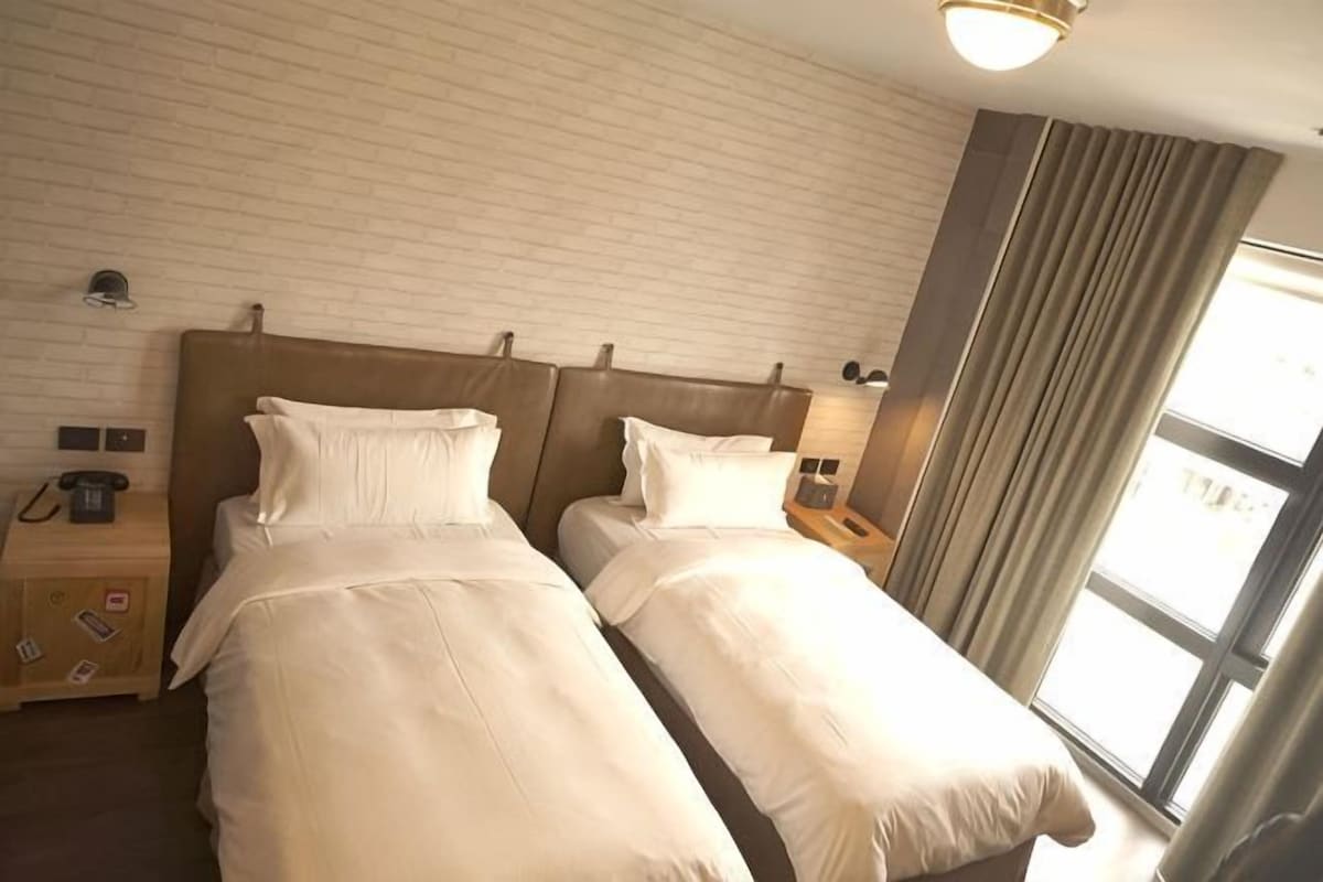 Ein Hotelzimmer mit zwei Betten, einer Wand mit Ziegeloptik, einem Fenster mit Vorhängen und Nachttischlampen.