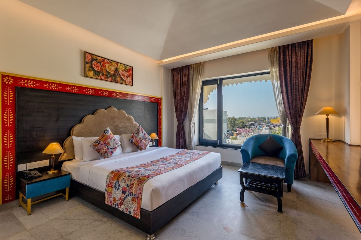 A hotel room with a large bed, a nightstand, a chair, a window with a view, and a painting on the wall.