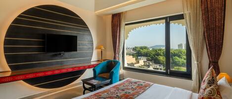 A hotel room with a bed, a TV mounted on a curved wall, a chair, and a window with curtains.