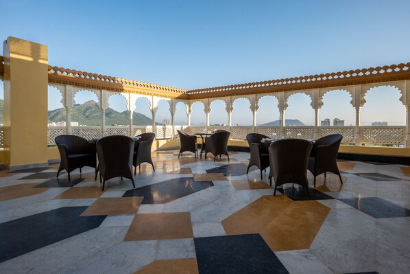 A terrace with a patterned floor, wicker furniture, and a view of distant hills. - Sterling Balicha Udaipur (Udaipur)
