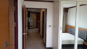 A hotel corridor with a wooden door on the left, a mirrored closet on the right, and a bed with a grey headboard in the background. - Ploenchit Superb Location, 86sqm, nearby Bumrungrad Hospital - minimum 30 days (Khwaeng Makkasan)