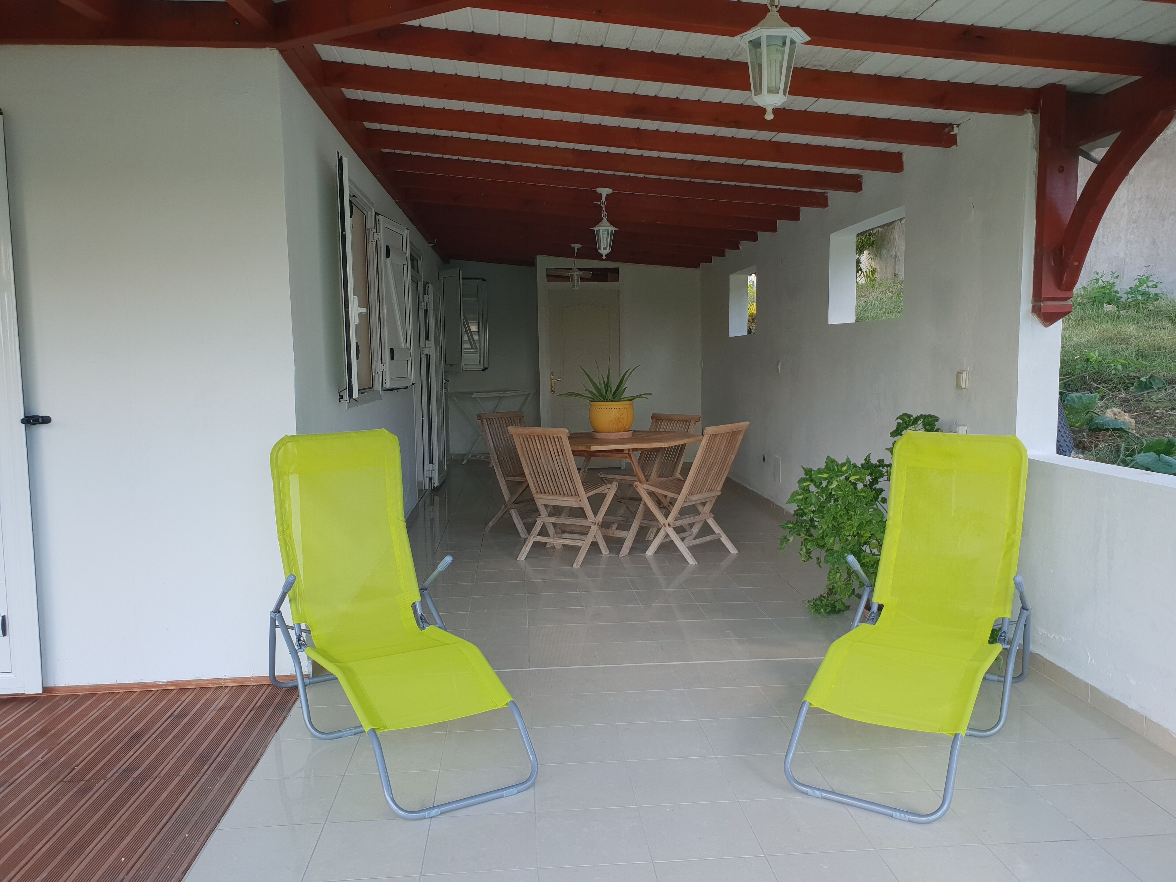 Een overdekt terras met een tafel en stoelen, een potplant en uitzicht op de omgeving.