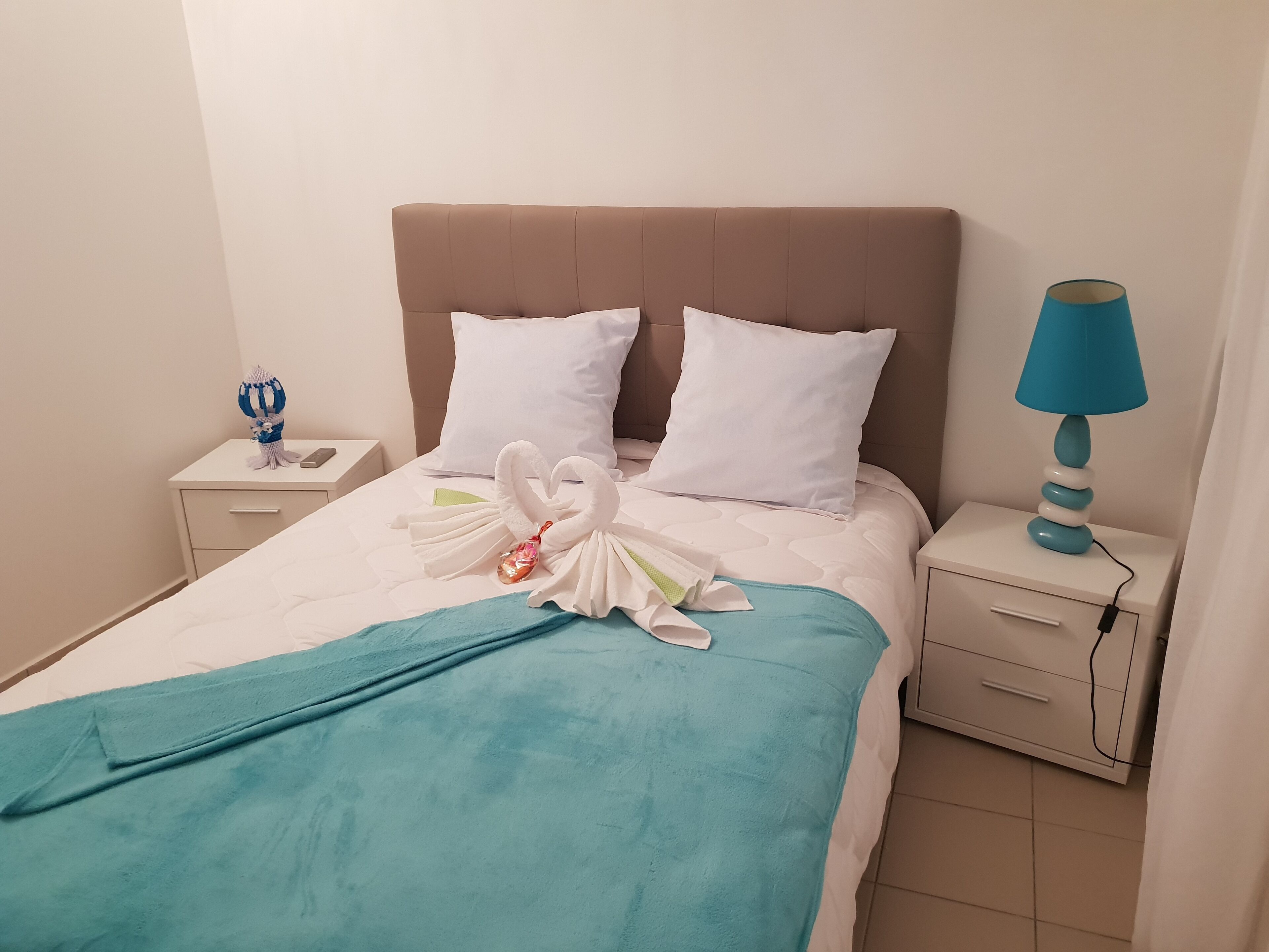 Ein ordentlich bezogenes Bett mit einer türkisblauen Decke, zwei weißen Kissen und einer zu einem Schwan geformten Handtuchdekoration. Auf dem Nachttisch steht eine blaue Lampe und ein kleines dekoratives Objekt.