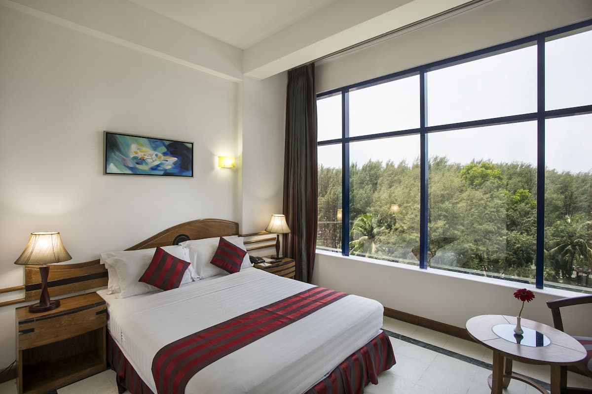 A hotel room with a large bed, two bedside tables with lamps, a small round table with a vase, a painting on the wall, and a view of trees through large windows.