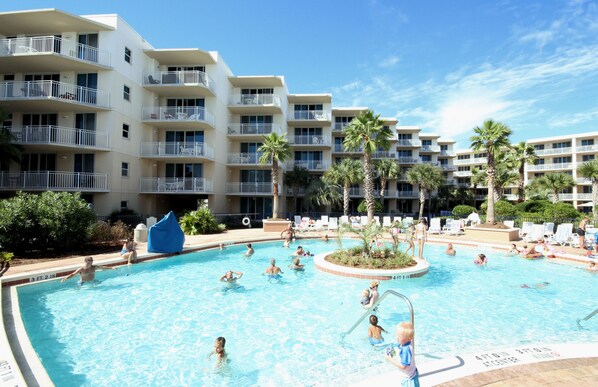 A swimming pool surrounded by multi-story buildings with balconies, palm trees, and lounge chairs.