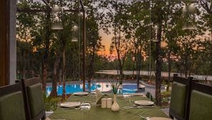 A dining table set for four with a green tablecloth, white plates, silverware, and a vase with greenery. The table is positioned near a window overlooking a pool and trees. - Siri Nature Roost (Chikkamagaluru)