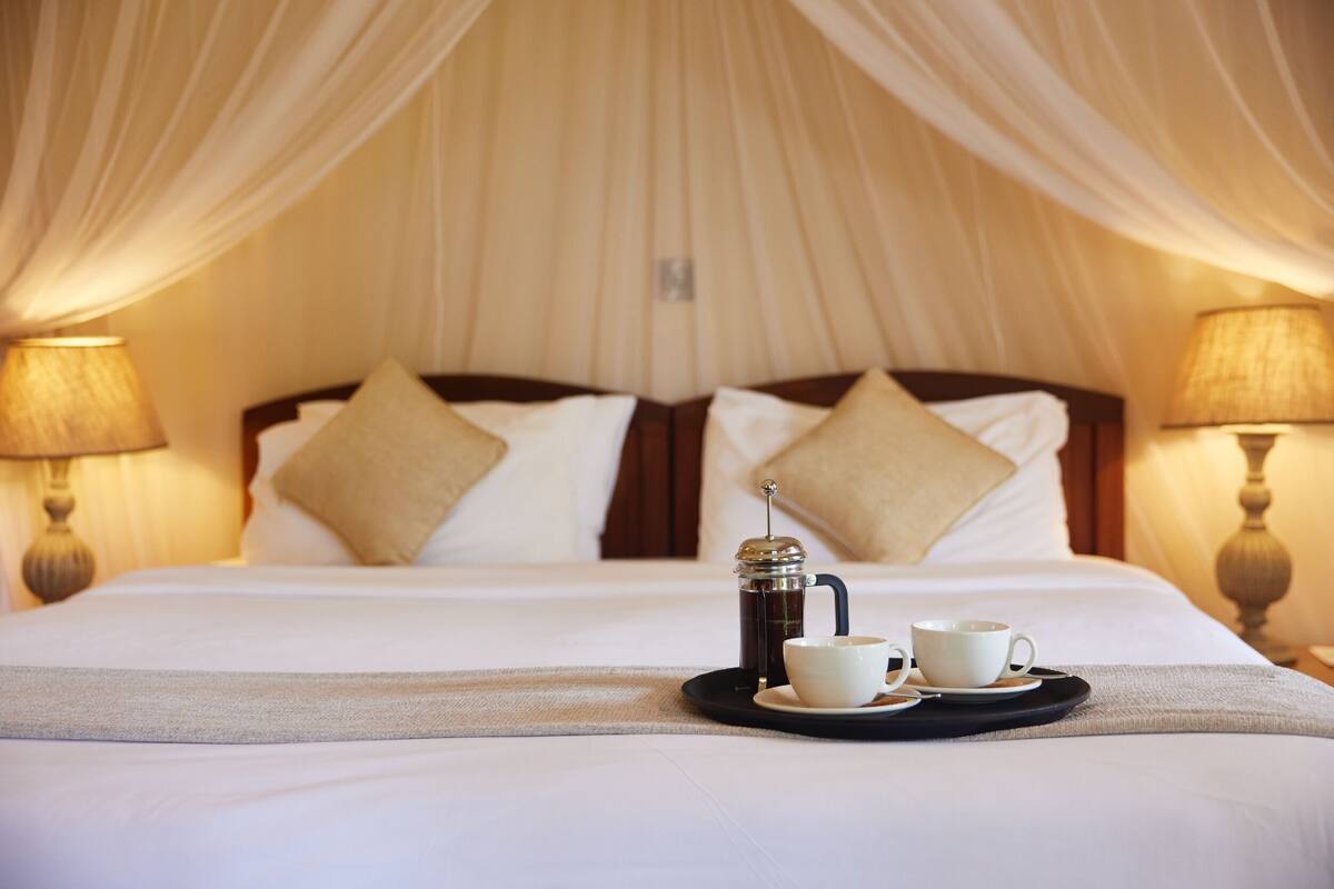 A bed with a canopy, two bedside lamps, a coffee maker, and two cups on a tray.