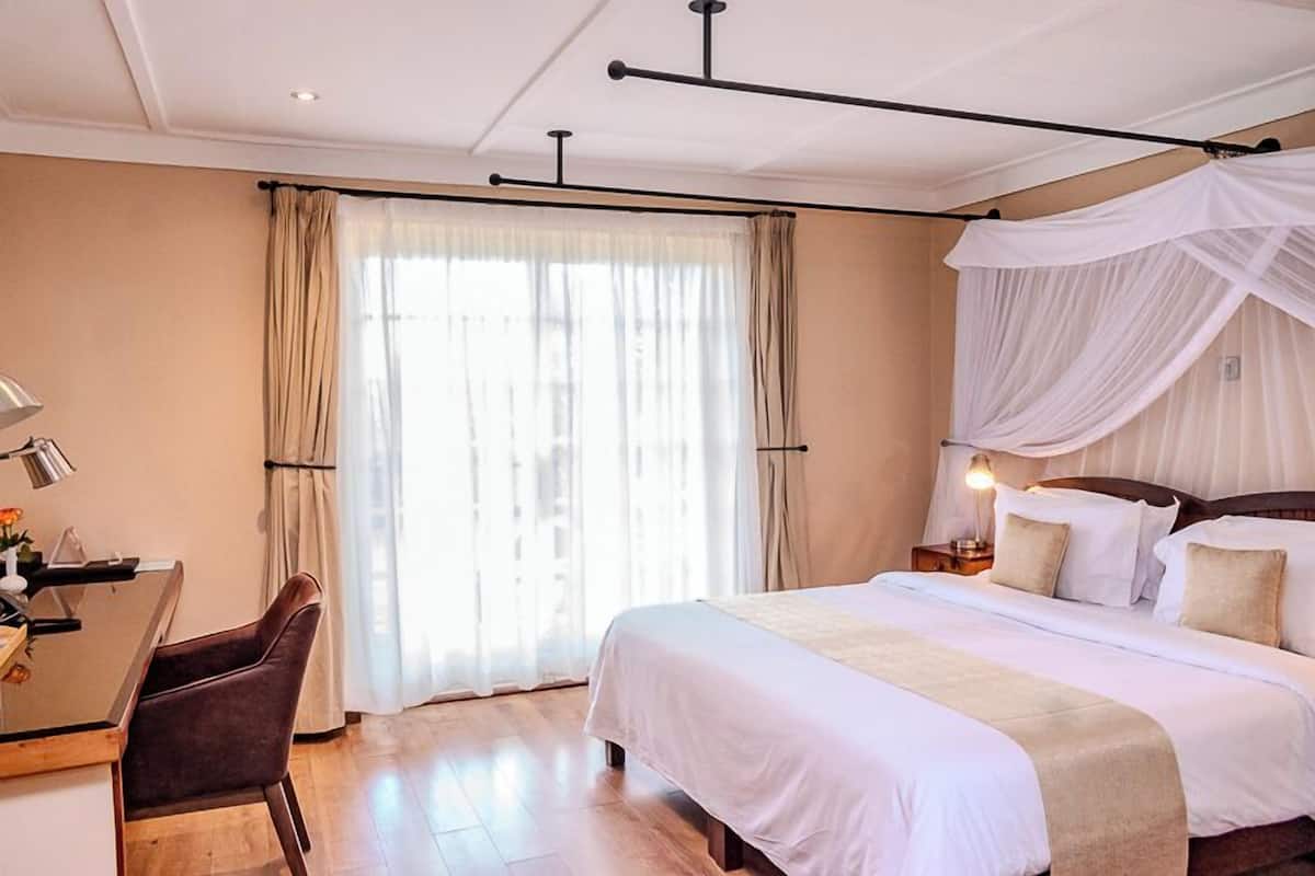 A hotel room with a bed, a desk with a chair, a bedside table with a lamp, and a large window with curtains.