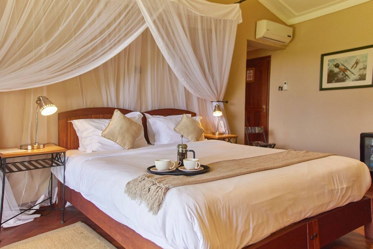A neatly made bed with a canopy, bedside lamps, a tray with cups and a coffee pot, and a framed picture on the wall.