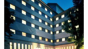 A multi-story hotel with a modern design, featuring a central entrance with a covered waiting area and a parking lot in front. - Aeria Tohno (Tono)