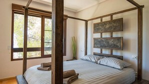 Una cama con dosel y vistas a árboles a través de amplias ventanas, una planta alta junto a la cama y un cabecero rústico de madera.