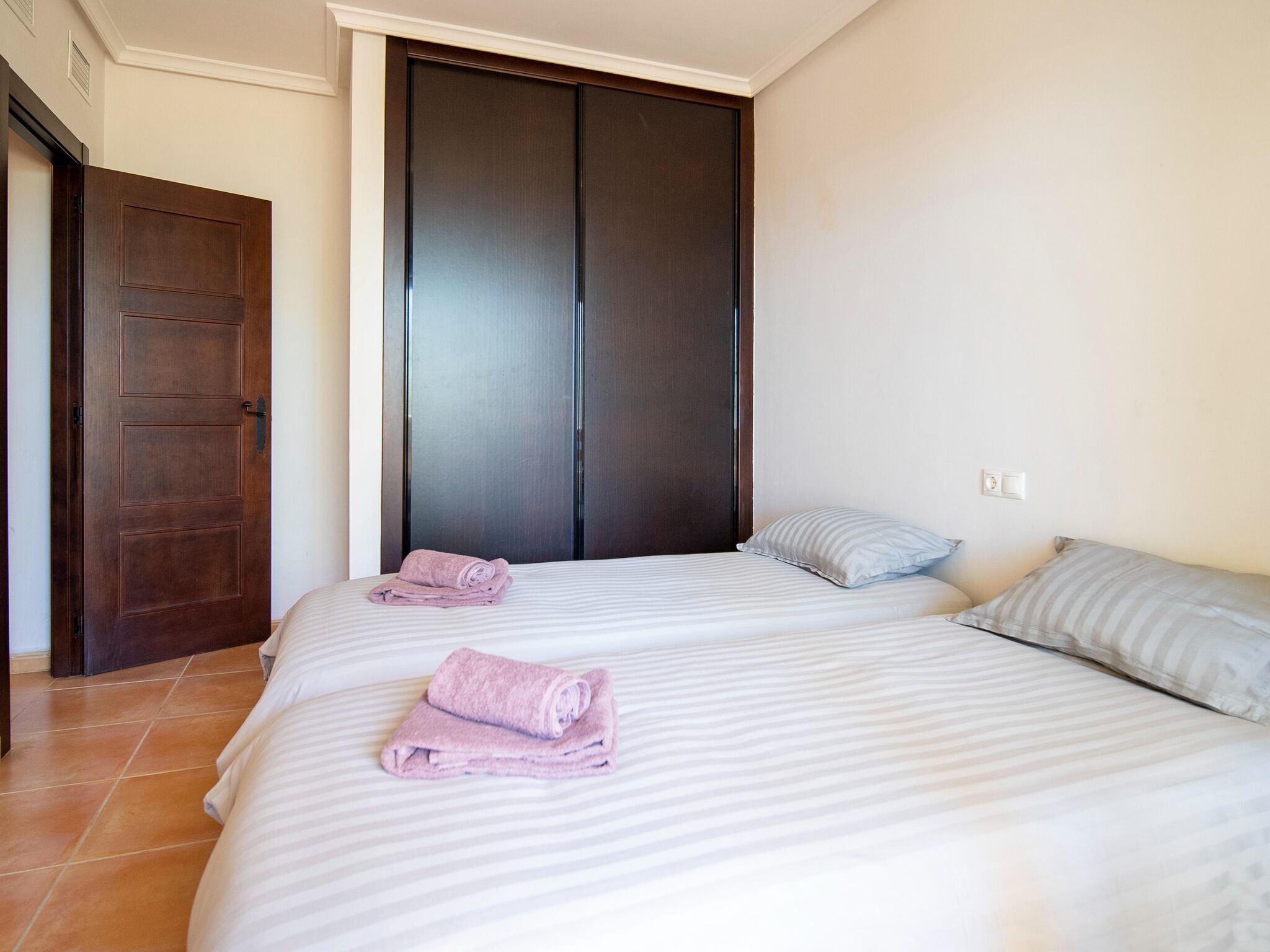 A bedroom with a large bed, two pillows, a folded towel, and a dark wooden wardrobe.