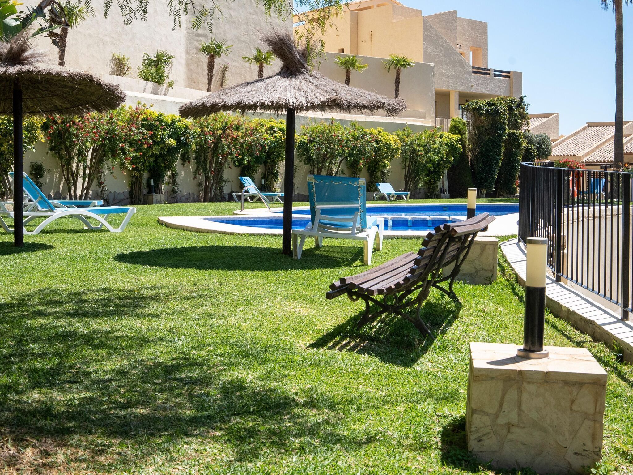 Zona de piscina con sillas de descanso y sombrillas, rodeada de vegetación y un edificio de techo plano.