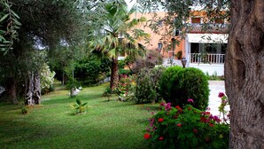 A lush garden with various plants and flowers, a well-maintained lawn, and a building with a white facade and red roof in the background. - Hotel Tina (Corfu)